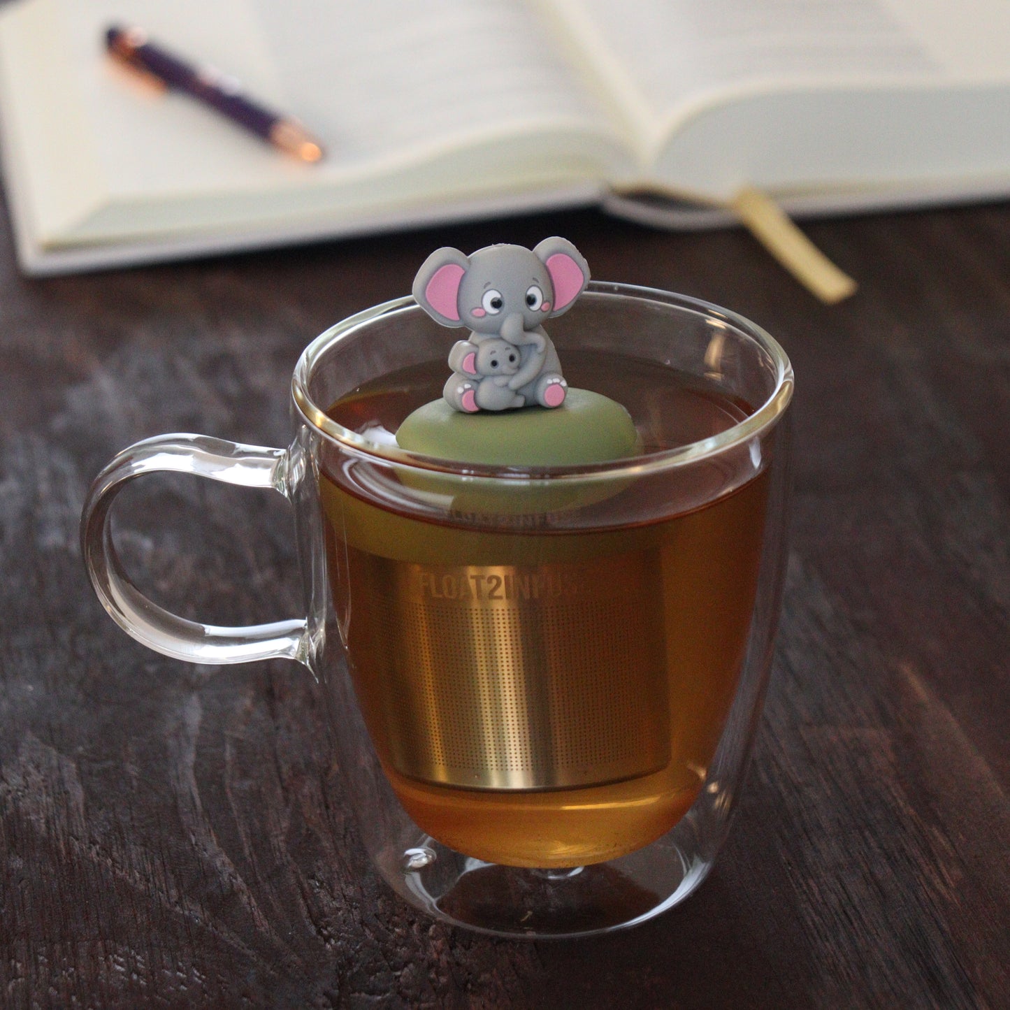 grey mom elephant and baby elephant animal tea infuser for loose leaf tea  with a fine mesh stainless steel tea infuser that reads FLOAT2INFUSE on a green base floating in a glass tea mug with notebook and pen in the background