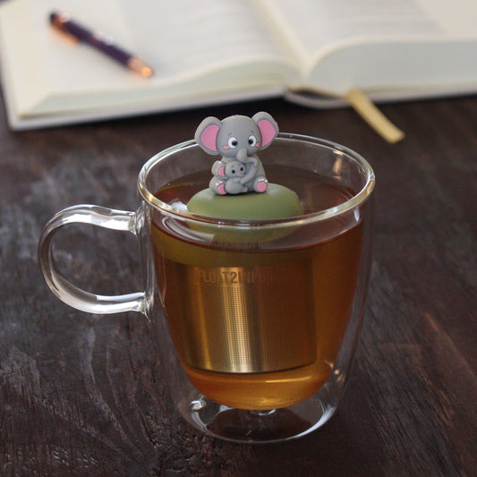 grey mom elephant and baby elephant animal tea infuser for loose leaf tea  with a fine mesh stainless steel tea infuser that reads FLOAT2INFUSE on a green base floating in a glass tea mug with notebook and pen in the background