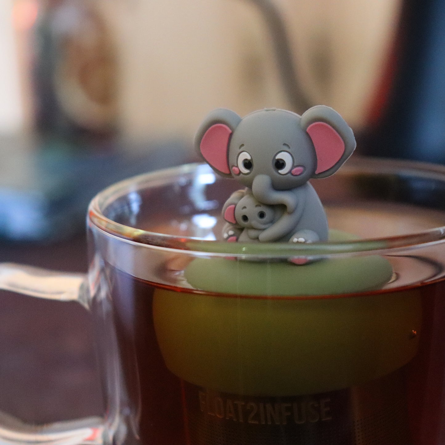 grey elephant mom and baby elephant animal tea infuser for loose leaf tea  with a fine mesh stainless steel tea strainer that reads FLOAT2INFUSE on a green base floating in a glass tea mug with tea kettle in the background