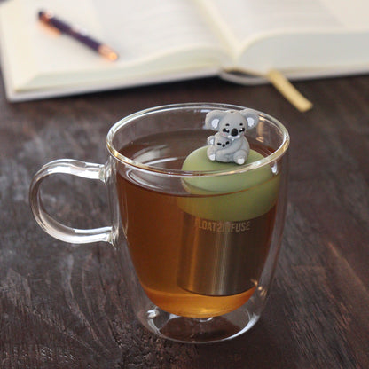 grey koala mom and baby koala animal tea infuser for loose leaf tea  with a fine mesh stainless steel tea infuser that reads FLOAT2INFUSE on a green base floating in a glass tea mug with notebook and pen in the background