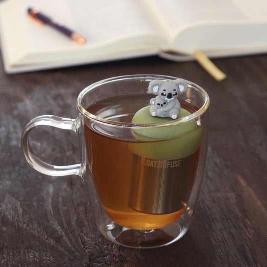 grey koala mom and baby koala animal tea infuser for loose leaf tea  with a fine mesh stainless steel tea infuser that reads FLOAT2INFUSE on a green base floating in a glass tea mug with notebook and pen in the background