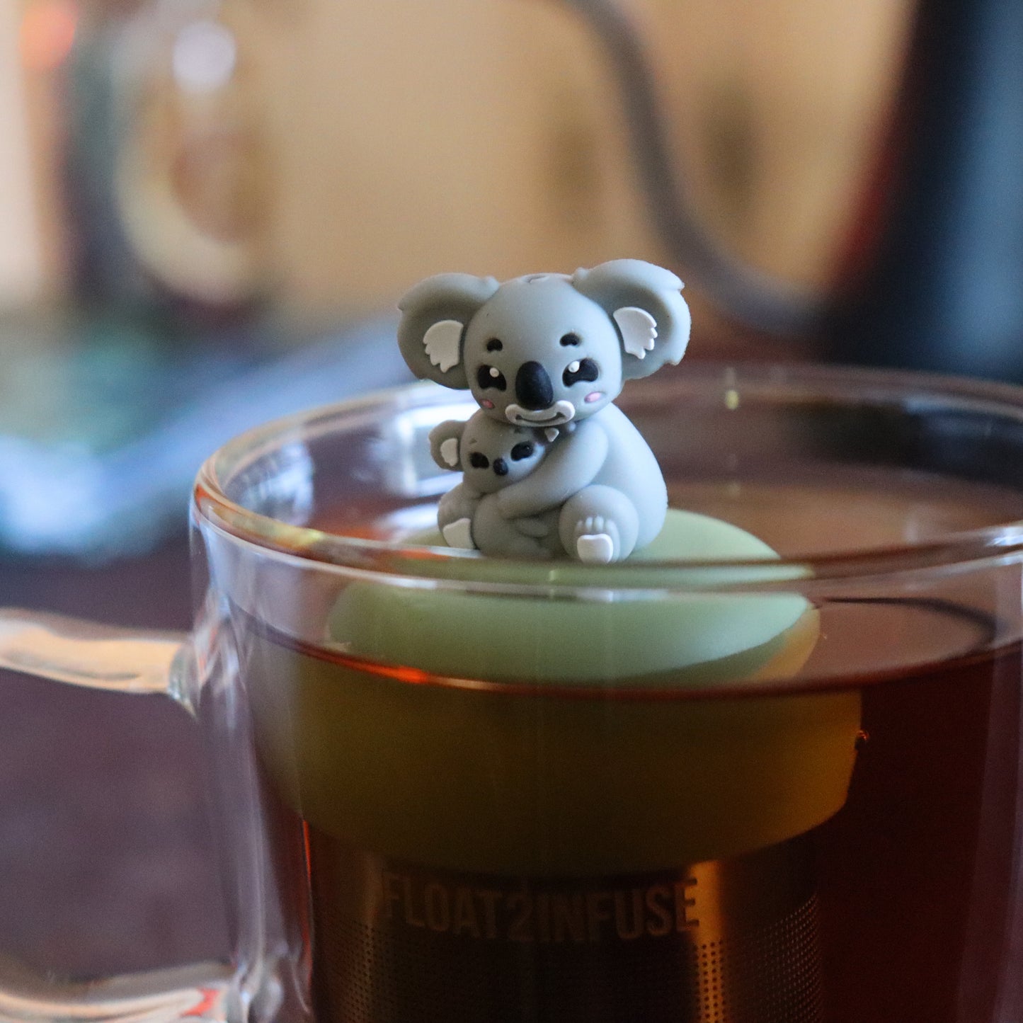 grey koala mom holding baby koala animal tea infuser for loose leaf tea  with a fine mesh stainless steel tea strainer that reads FLOAT2INFUSE on a green base floating in a glass tea mug with tea kettle in the background