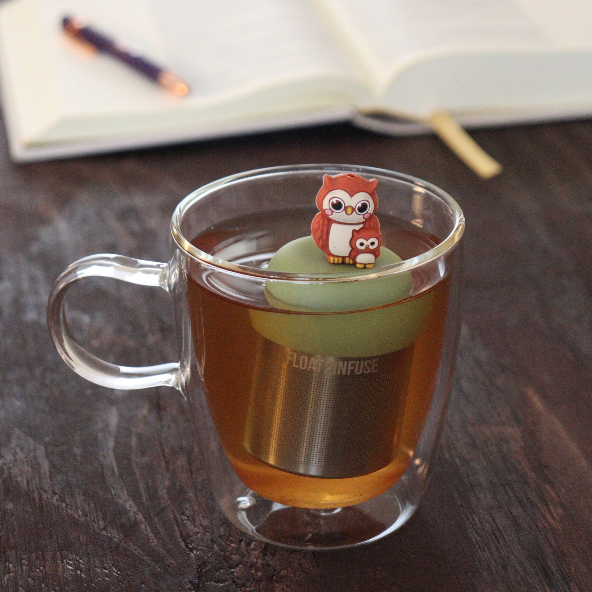 brown mom and baby owl animal tea infuser for loose leaf tea  with a fine mesh stainless steel tea infuser that reads FLOAT2INFUSE on a green base floating in a glass tea mug with notebook and pen in the background