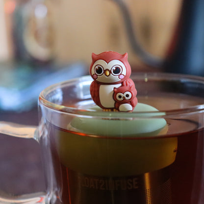 brown mom and baby owl animal tea infuser for loose leaf tea with a fine mesh stainless steel tea strainer that reads FLOAT2INFUSE on a green base  floating in a glass tea mug with tea kettle in the background