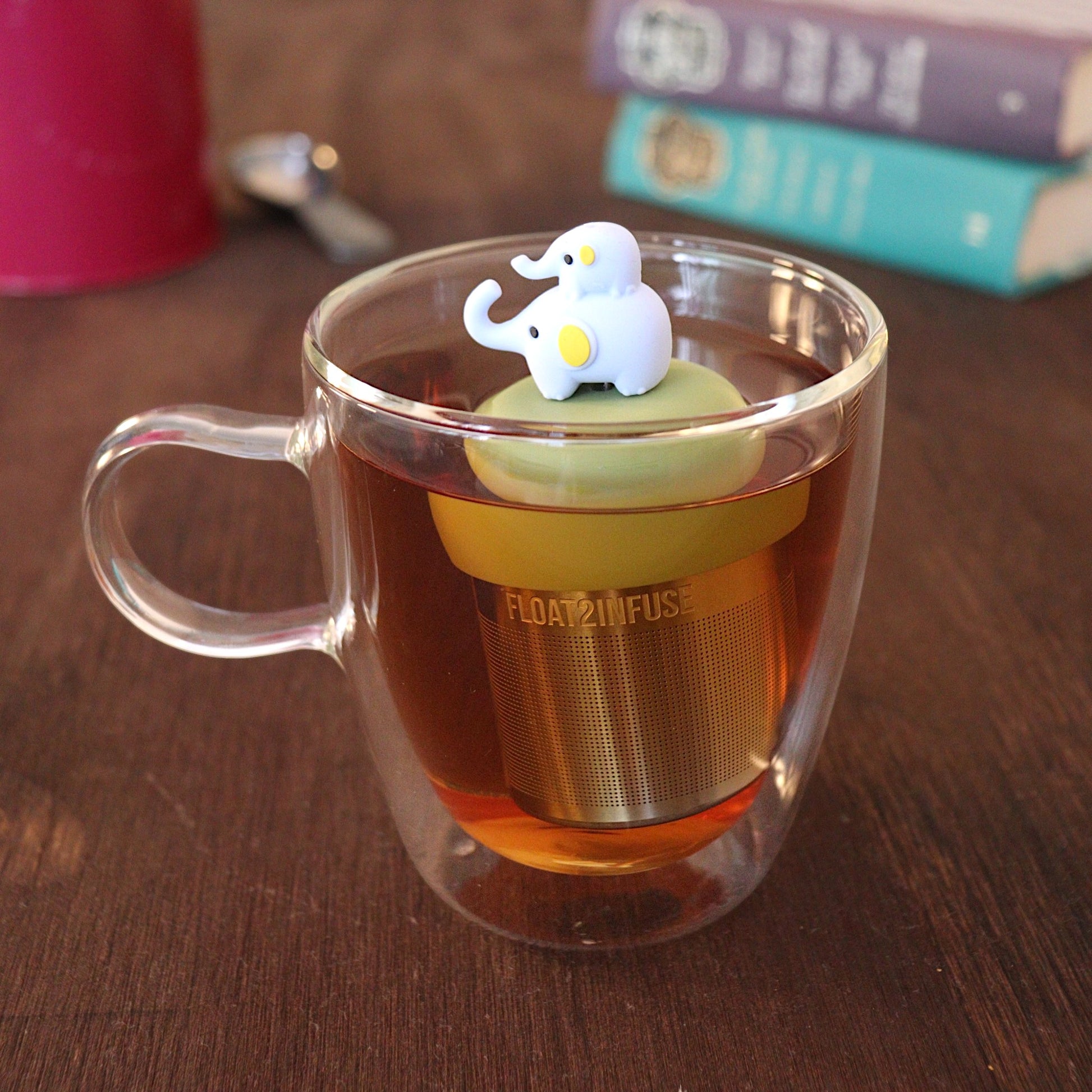Momma elephant with baby elephant riding on her back tea infuser for loose leaf tea with a fine mesh stainless steel tea strainer 
that reads FLOAT2INFUSE on a green base with a tea canister and books in the background