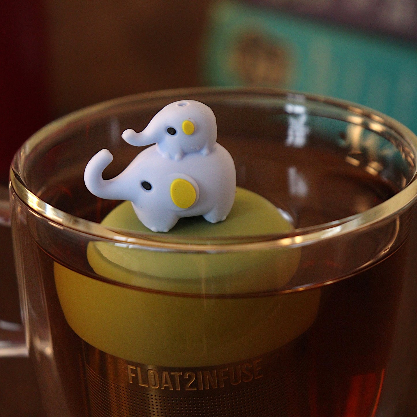 Momma elephant with baby elephant riding on her back tea infuser for loose leaf tea with a fine mesh stainless steel tea strainer 
that reads FLOAT2INFUSE on a green base with a tea canister and books in the background