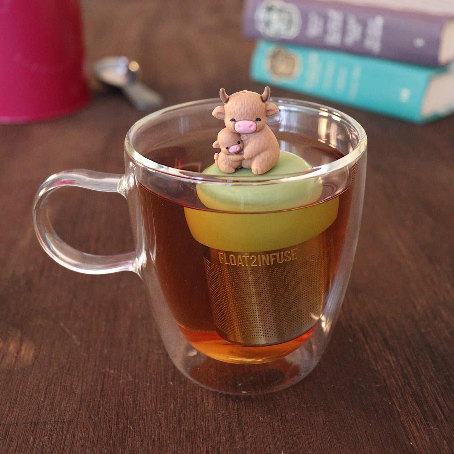 Momma highland cow hugging her baby tea infuser for loose leaf tea with a fine mesh stainless steel tea strainer that reads FLOAT2INFUSE on a green base with a tea canister and books in the backgroundåç