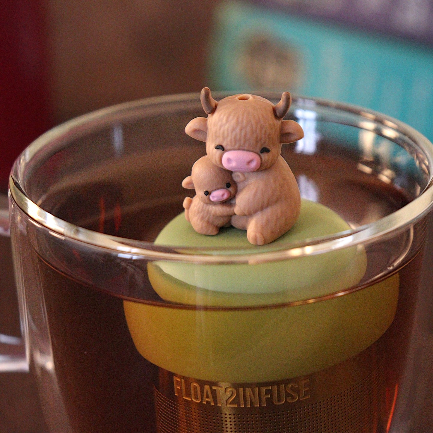 Momma highland cow hugging her baby tea infuser for loose leaf tea with a fine mesh stainless steel tea strainer that reads FLOAT2INFUSE on a green base with a tea canister and books in the backgroundåç