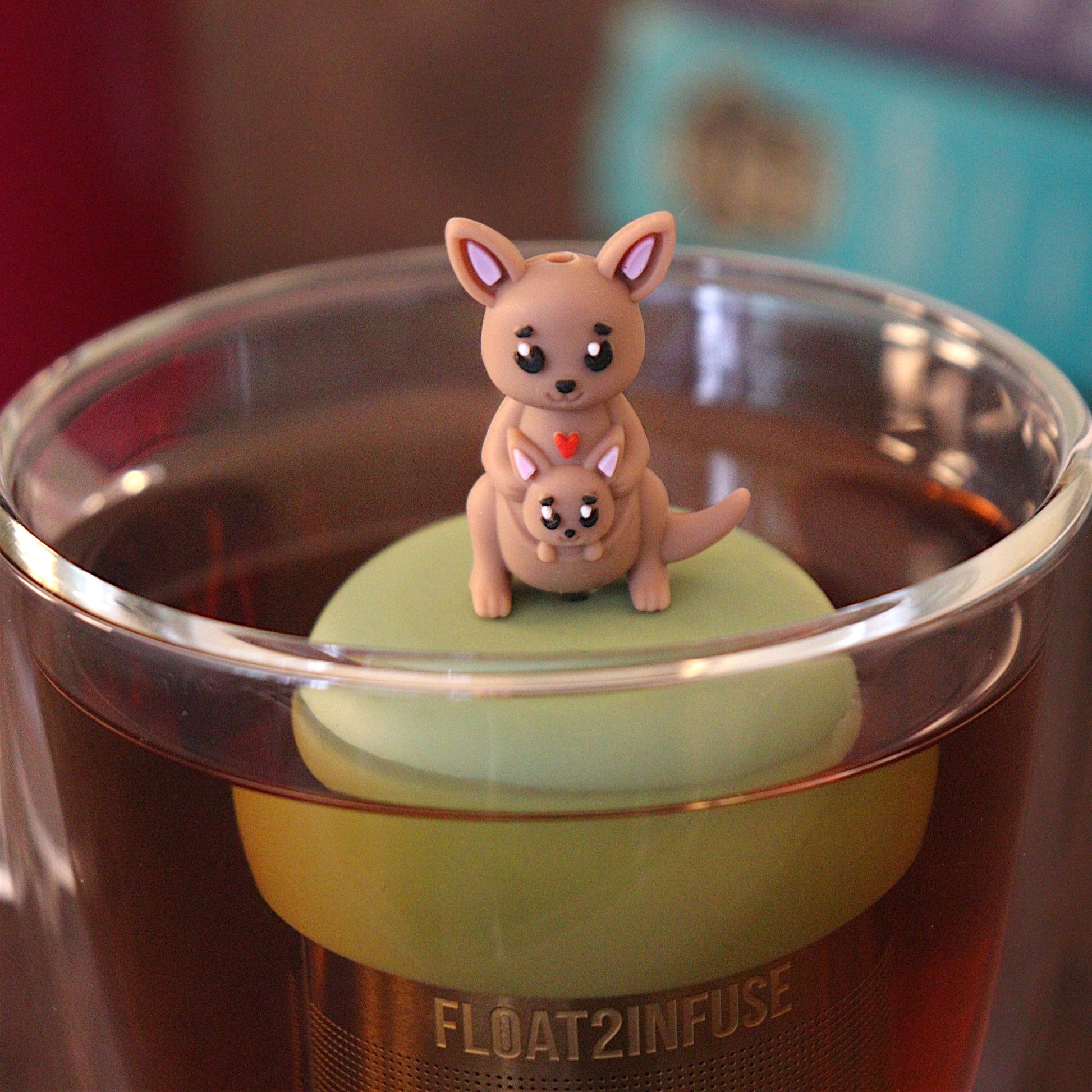 Cute mother kangaroo with a baby joey kangaroo in her pouch tea infuser for loose leaf tea with a fine mesh stainless steel tea strainer 
that reads FLOAT2INFUSE on a green base with a tea canister and books in the background