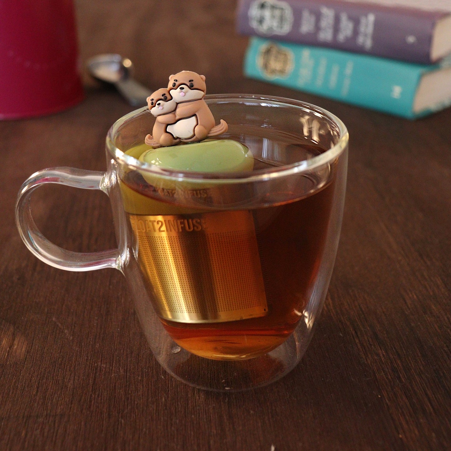 Brown otter momma hugging her baby tea infuser for loose leaf tea with a fine mesh stainless steel tea strainer that reads FLOAT2INFUSE on a green base with a tea canister and books in the background
