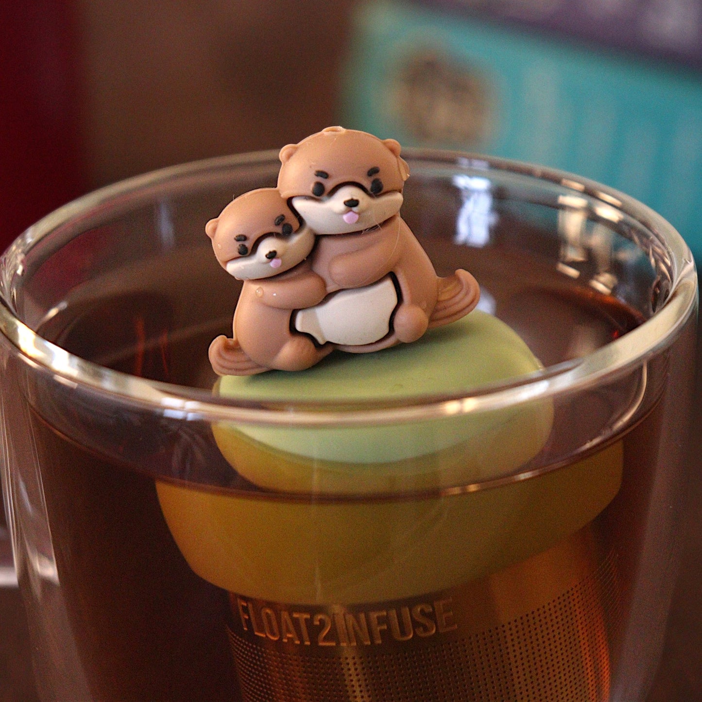 Brown otter momma hugging her baby tea infuser for loose leaf tea with a fine mesh stainless steel tea strainer that reads FLOAT2INFUSE on a green base with a tea canister and books in the background
