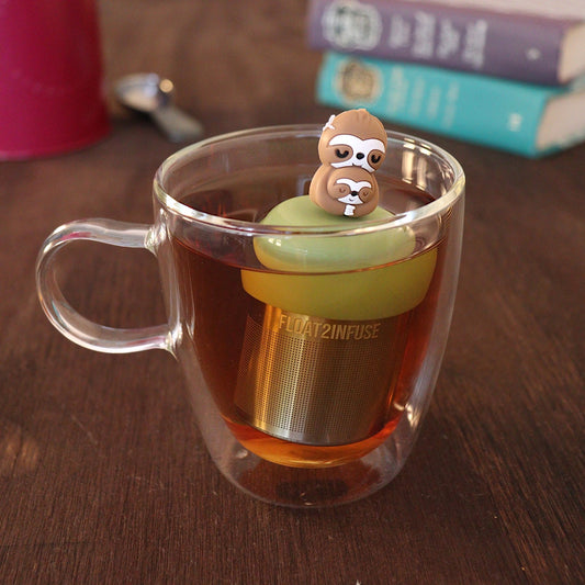Mom and baby sloth tea infuser for loose leaf tea with a fine mesh stainless steel tea strainer 
that reads FLOAT2INFUSE on a green base floating in a glass tea mug 
with a tea canister and books in the background
