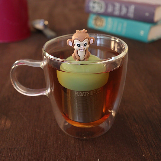 Smiling brown monkey tea infuser for loose leaf tea with a fine mesh stainless steel tea strainer 
that reads FLOAT2INFUSE on a green base with a tea canister and books in the background
