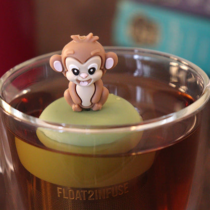 Smiling brown monkey tea infuser for loose leaf tea with a fine mesh stainless steel tea strainer 
that reads FLOAT2INFUSE on a green base with a tea canister and books in the background