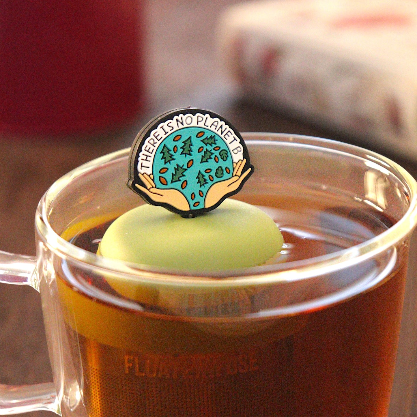 black circle with open hands holding images of leaves and pine trees, and text that reads "There is no planet b"  tea infuser for loose leaf tea on a green base with a fine mesh stainless steel tea infuser that reads FLOAT2INFUSE floating in a glass tea mug with pen on notebook and loose leaf tea and books in the background