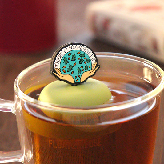 black circle with open hands holding images of leaves and pine trees, and text that reads "There is no planet b"  tea infuser for loose leaf tea on a green base with a fine mesh stainless steel tea infuser that reads FLOAT2INFUSE floating in a glass tea mug with pen on notebook and loose leaf tea and books in the background