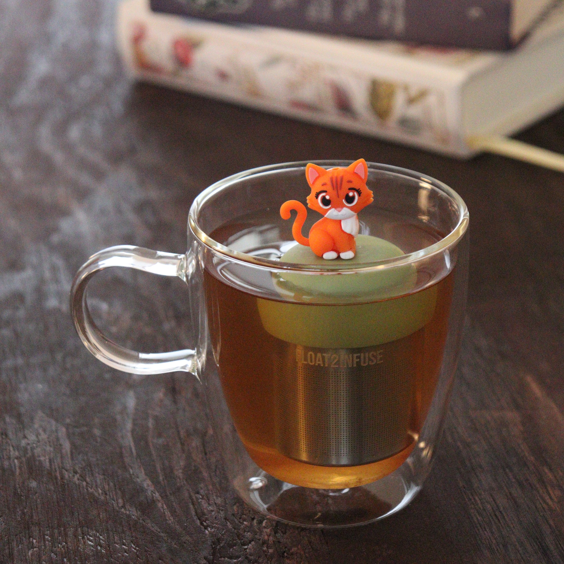 orange cat animal tea infuser for loose leaf tea with a fine mesh stainless steel tea infuser that reads FLOAT2INFUSE on a green base  floating in a glass tea mug with books in the background