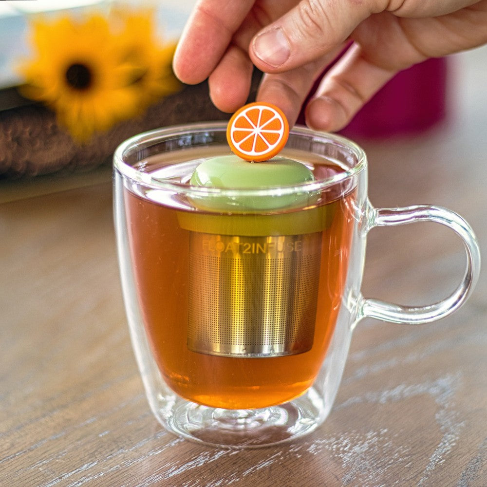 orange fruit slice tea infuser for loose leaf tea  with a fine mesh stainless steel tea infuser that reads FLOAT2INFUSE on a green base floating in a glass tea mug with flowers in the background
