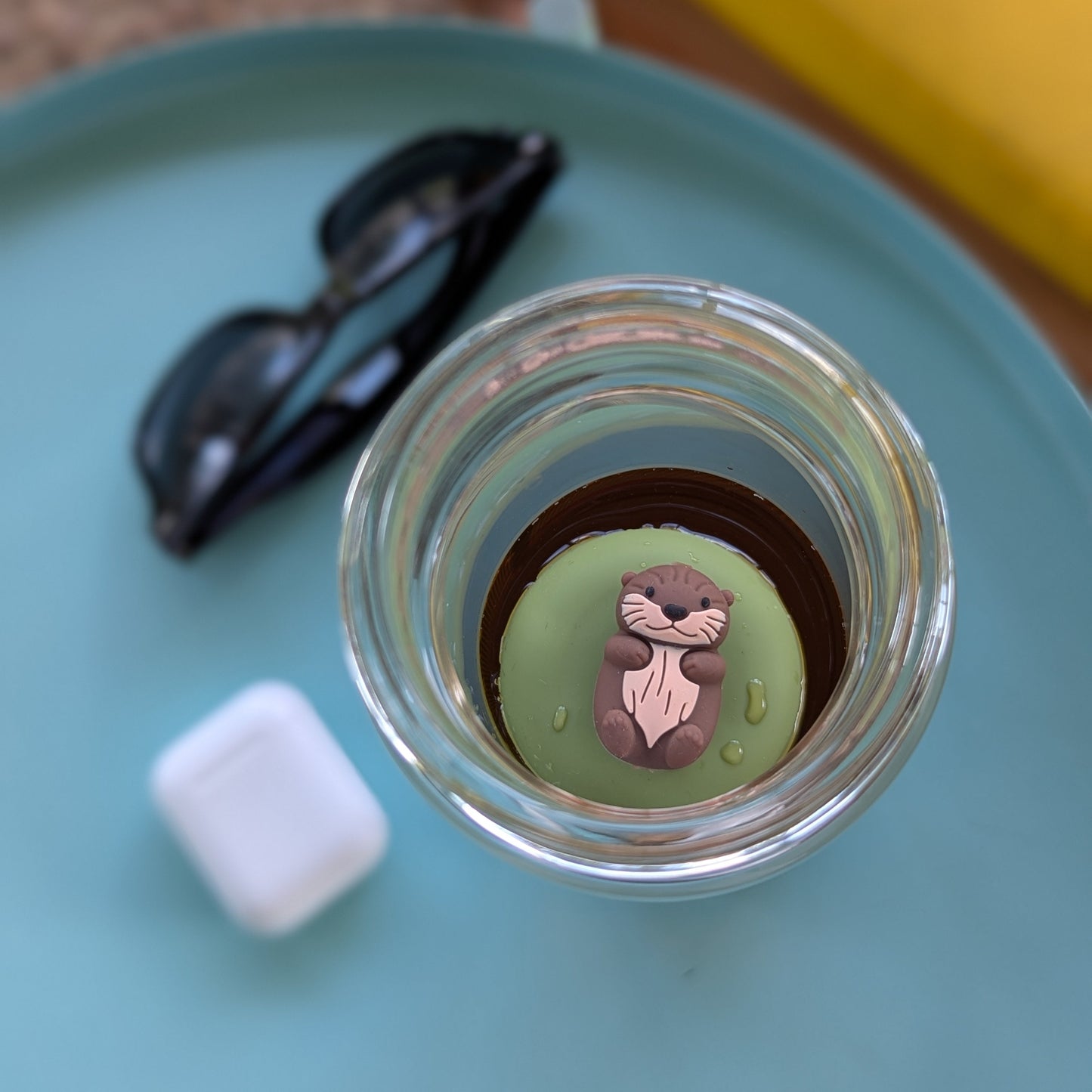 top view of a brown otter tea infuser for loose leaf tea with a fine mesh stainless steel tea strainer that reads FLOAT2INFUSE on a green base floating in a glass tea mug with sunglasses and earbuds in the background
