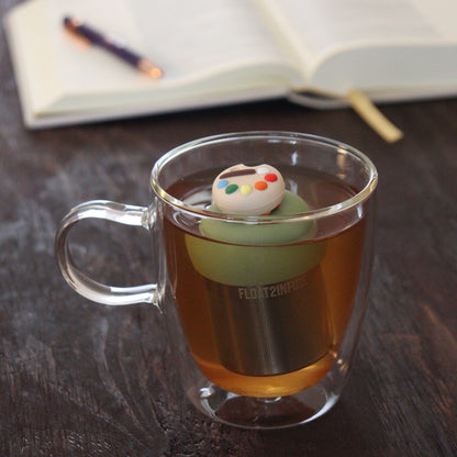 paint palette tea infuser for loose leaf tea with a fine mesh stainless steel tea infuser that reads FLOAT2INFUSE on a green base floating in a glass tea mug with notebook and pen in the background