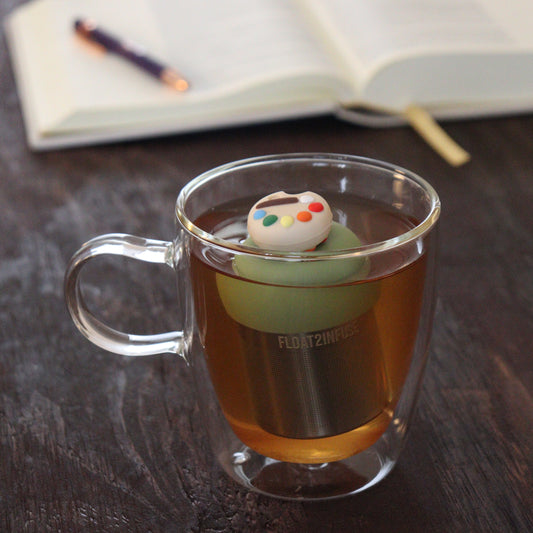 paint palette tea infuser for loose leaf tea with a fine mesh stainless steel tea infuser that reads FLOAT2INFUSE on a green base floating in a glass tea mug with notebook and pen in the background