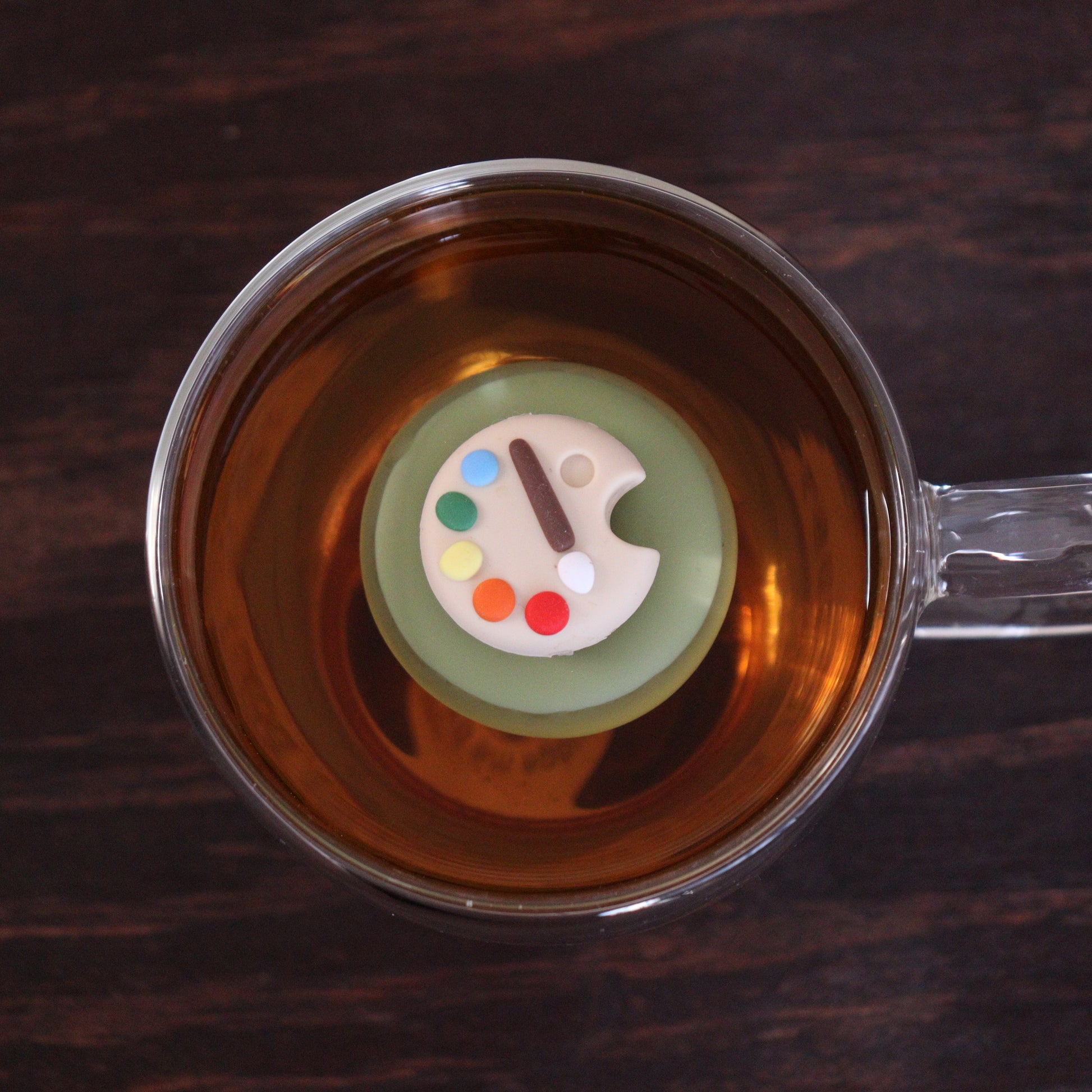 paint palette tea infuser for loose leaf tea  with a fine mesh stainless steel tea infuser that reads FLOAT2INFUSE on a green base floating in a glass tea mug sitting on a wooden table