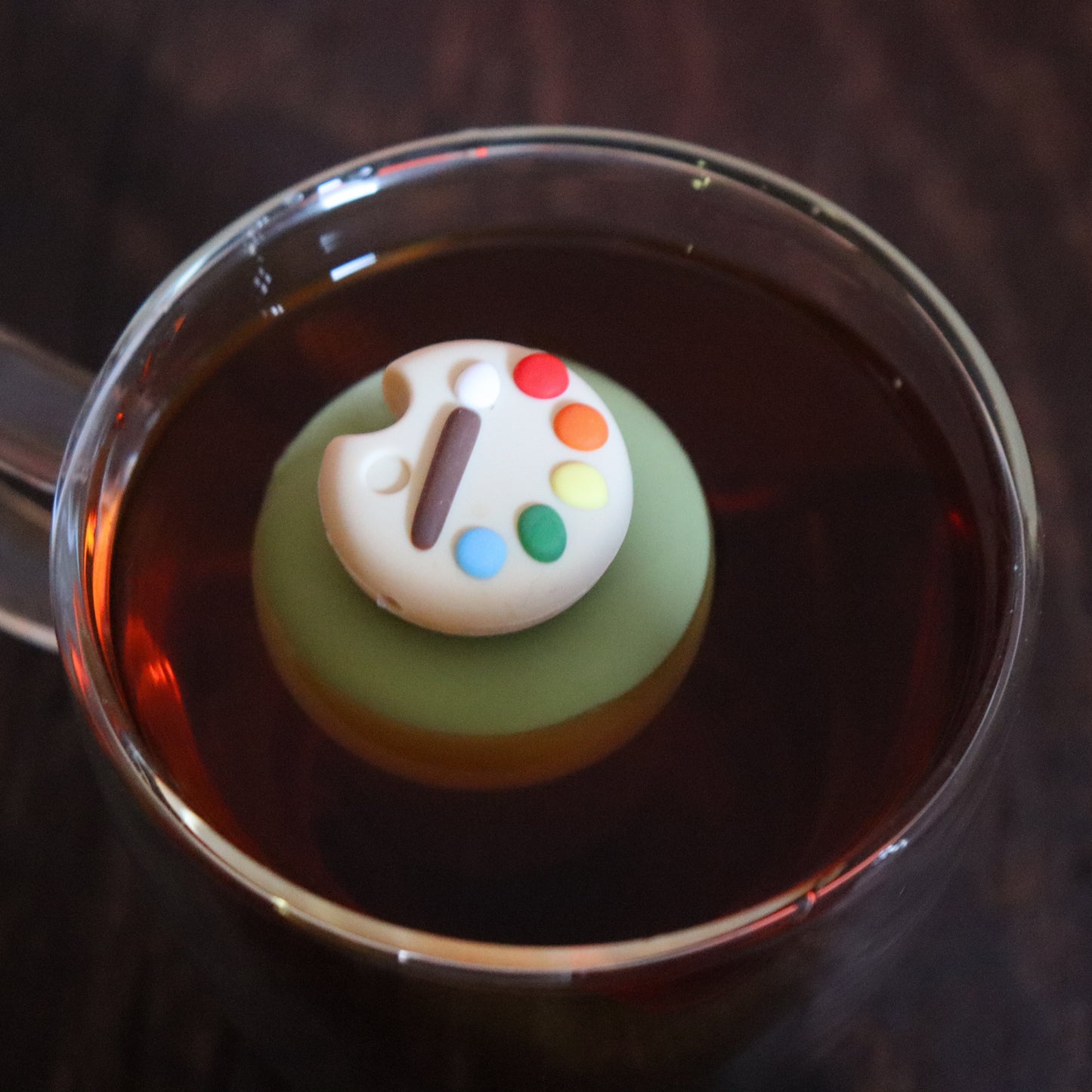 paint palette tea infuser for loose leaf tea  with a fine mesh stainless steel tea strainer that reads FLOAT2INFUSE on a green base floating in a glass tea mug with tea kettle in the background