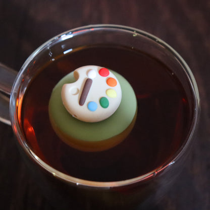 paint palette tea infuser for loose leaf tea  with a fine mesh stainless steel tea strainer that reads FLOAT2INFUSE on a green base floating in a glass tea mug with tea kettle in the background
