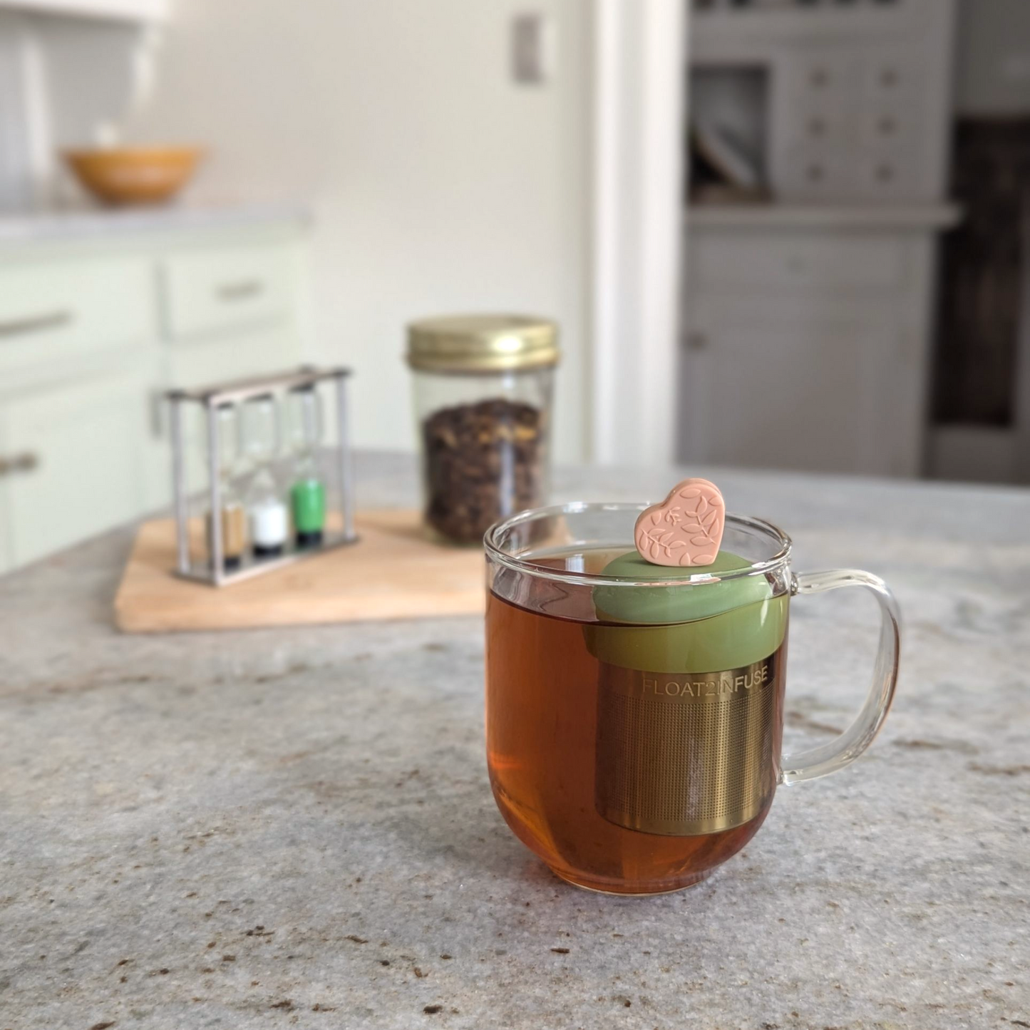 peach heart with a floral leaf pattern tea infuser  for loose leaf tea with fine mesh stainless steel tea strainer that reads FLOAT2INFUSE on a green base floating in a glass tea mug with loose leaf tea in the background