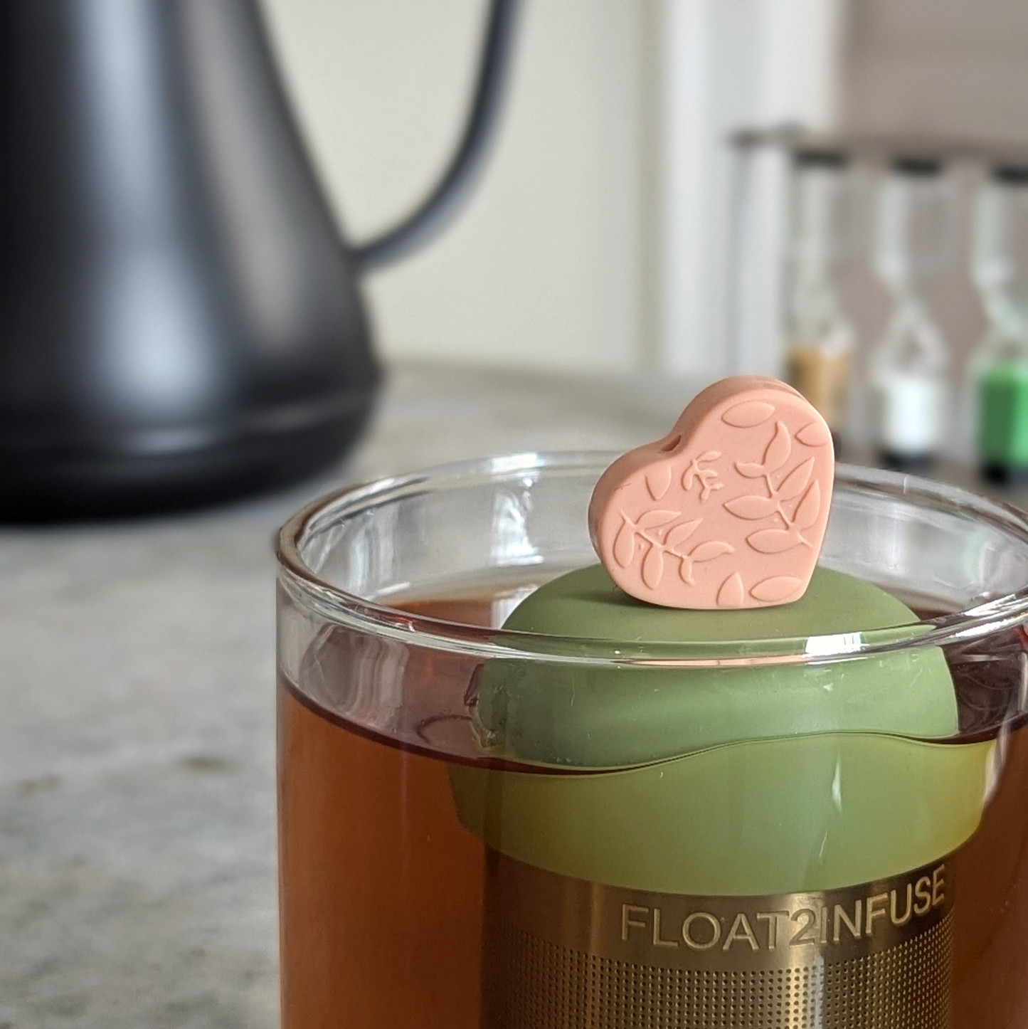peach heart with a floral leaf pattern tea infuser  for loose leaf tea with fine mesh stainless steel tea strainer that reads FLOAT2INFUSE on a green base floating in a glass tea mug with a tea kettle in the background