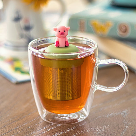 pink pig tea infuser for loose leaf tea with a fine mesh stainless steel tea strainer that reads FLOAT2INFUSE on a green base  floating in a glass tea mug 
with books in the background