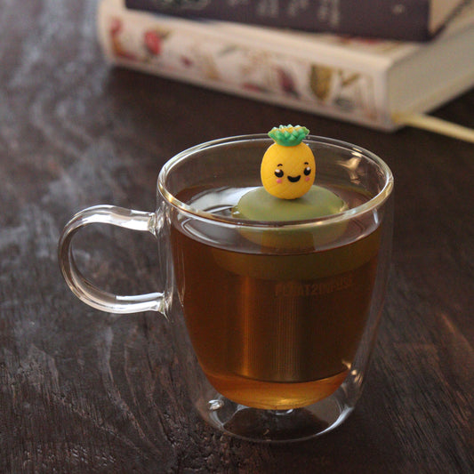 pineapple tea infuser for loose leaf tea on a green base with a fine mesh stainless steel tea infuser that reads FLOAT2INFUSE floating in a glass tea mug with books in the background