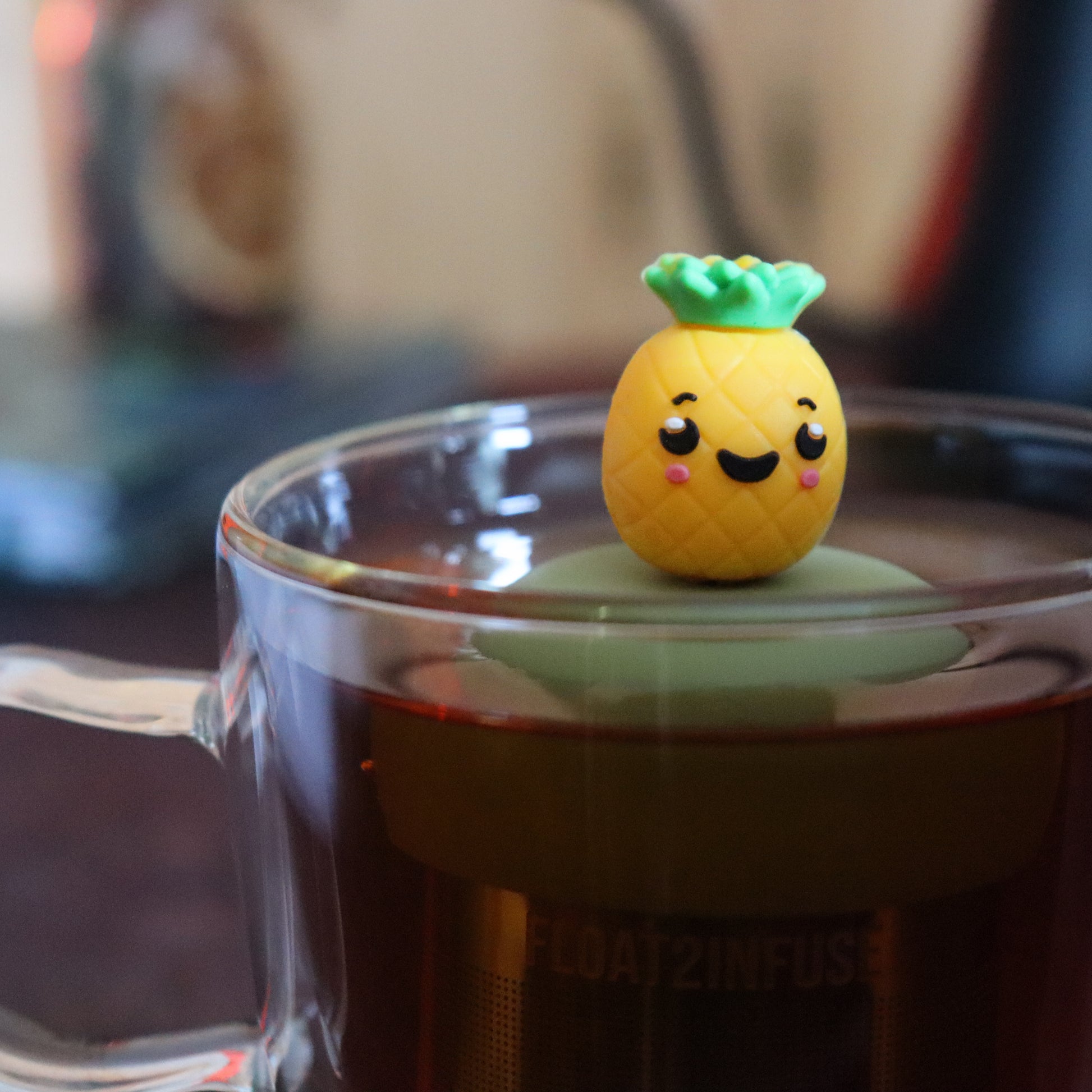 cute pineapple tea infuser for loose leaf tea on a green base with a fine mesh stainless steel tea strainer that reads FLOAT2INFUSE floating in a glass tea mug with tea kettle in the background