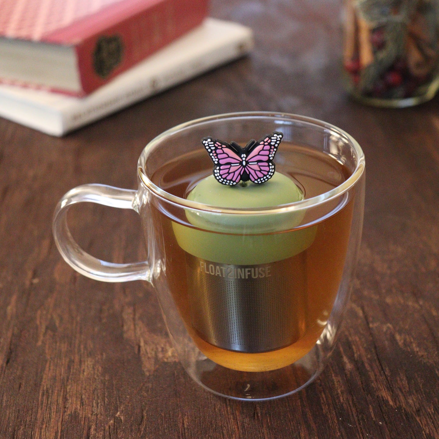 pink animal tea infuser for loose leaf tea with a fine mesh stainless steel tea infuser that reads FLOAT2INFUSE on a green base floating in a glass tea mug with books and loose leaf tea in the background