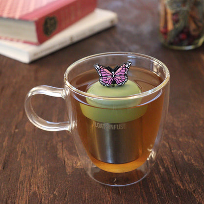 pink animal tea infuser for loose leaf tea with a fine mesh stainless steel tea infuser that reads FLOAT2INFUSE on a green base floating in a glass tea mug with books and loose leaf tea in the background