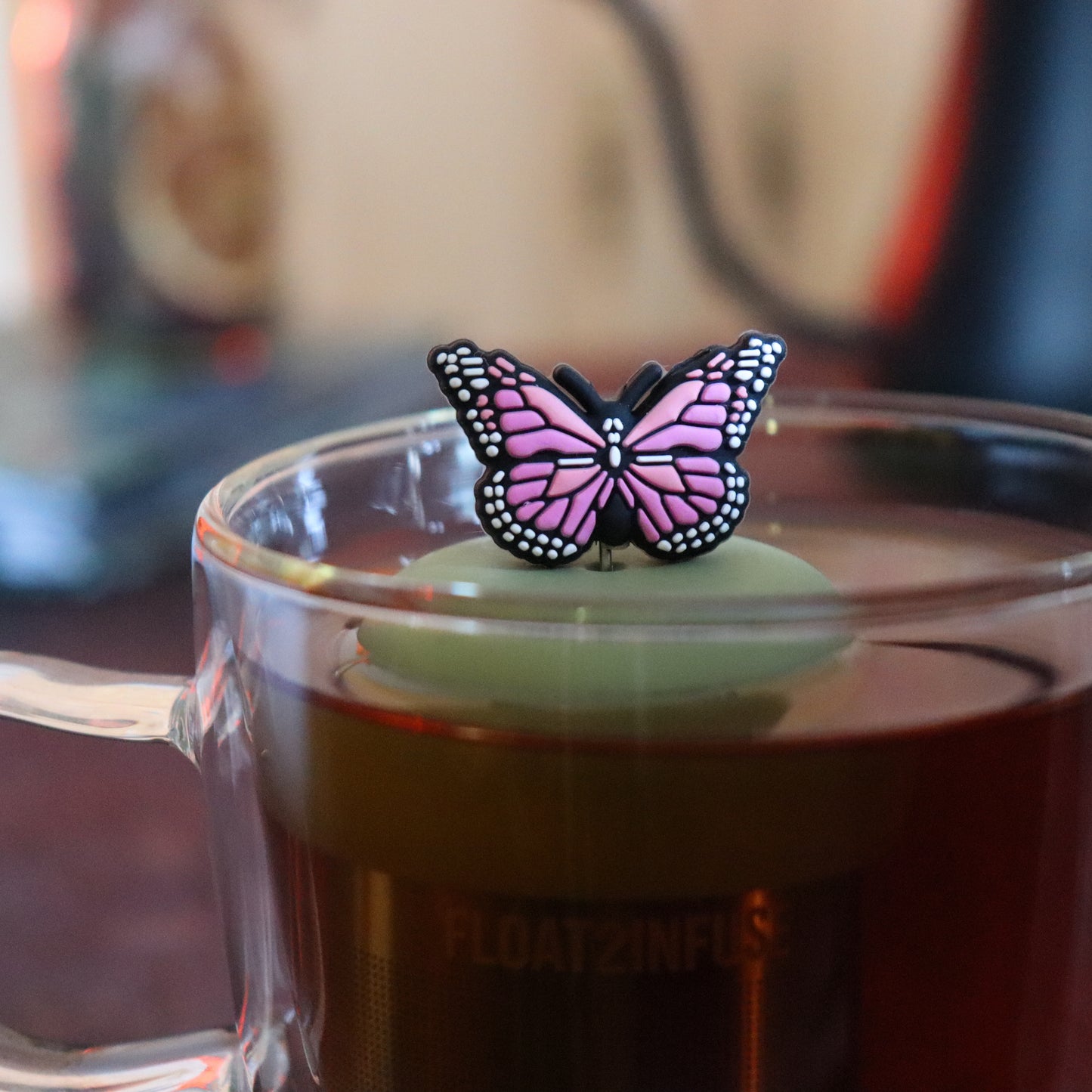pink butterfly tea infuser for loose leaf tea  with a fine mesh stainless steel tea strainer that reads FLOAT2INFUSE on a green base floating in a glass tea mug with tea kettle in the background