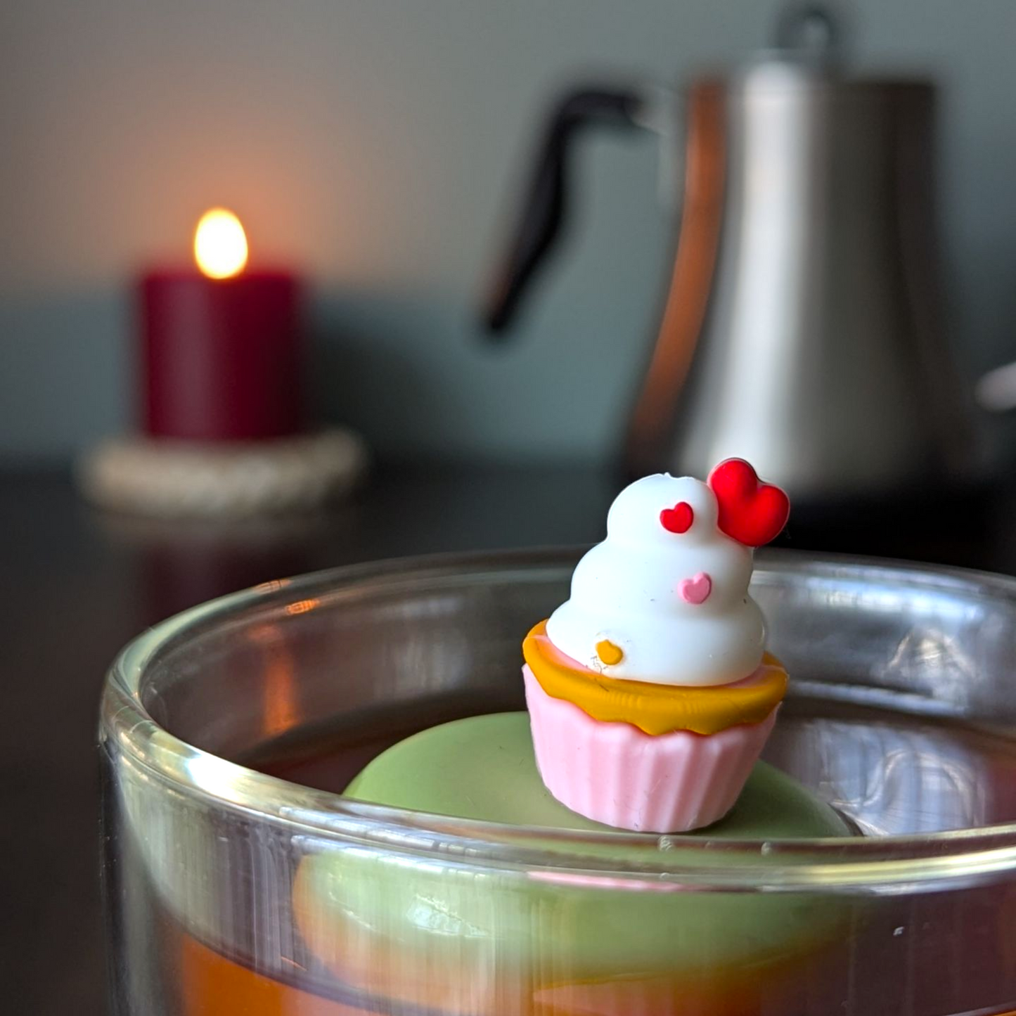 vanilla cupcake in a pink wrapper with white icing and heart shaped sprinkles tea infuser for loose leaf tea with a fine mesh stainless steel tea strainer that reads FLOAT2INFUSE on a green base  with a lit candle and a tea kettle in the background