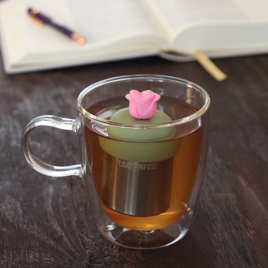 pink rose tea infuser for loose leaf tea with a fine mesh stainless steel tea infuser that reads FLOAT2INFUSE on a green base floating in a glass tea mug with notebook and pen in the background