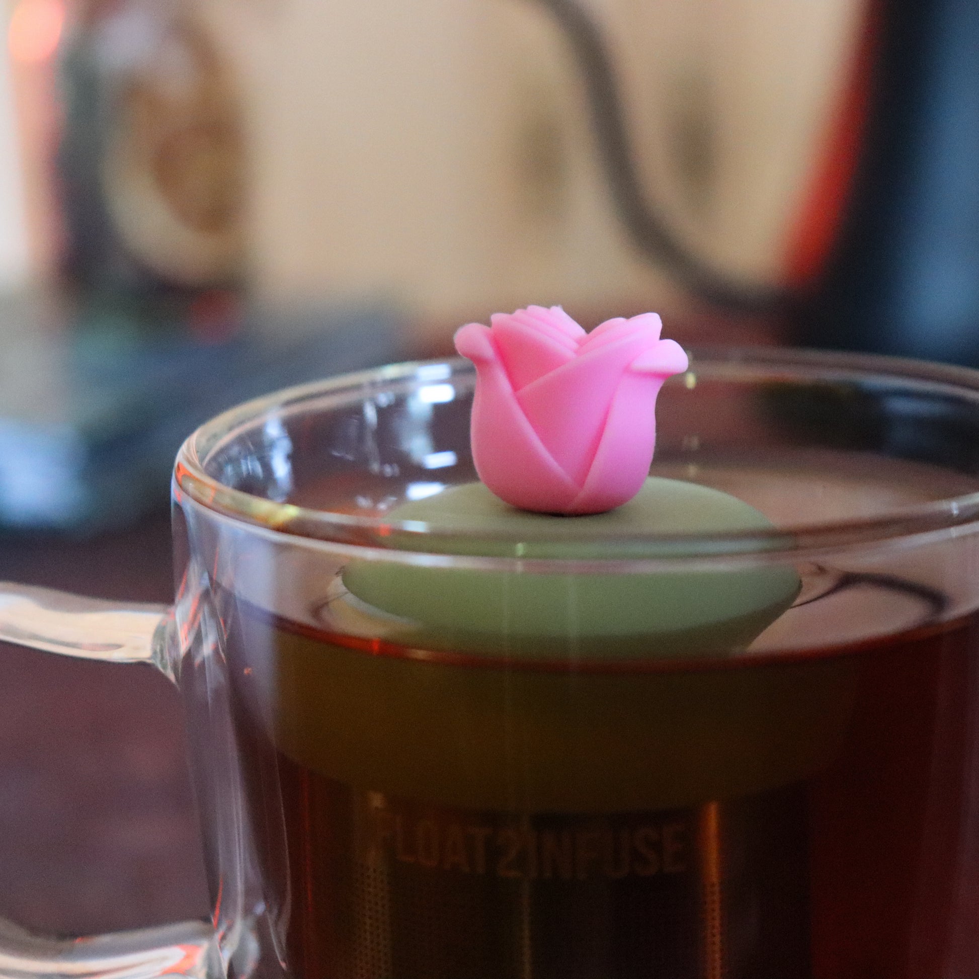 pink rose tea infuser for loose leaf tea  with a fine mesh stainless steel tea strainer that reads FLOAT2INFUSE on a green base floating in a glass tea mug with tea kettle in the background