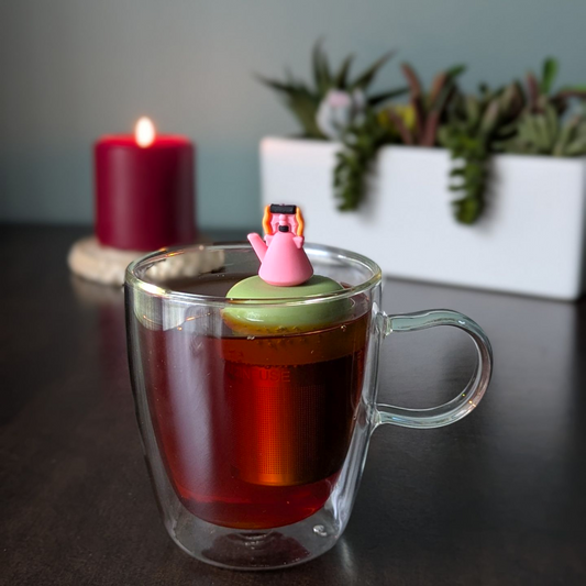 pink tea kettle with orange and black handle tea infuser  for loose leaf tea with fine mesh stainless steel tea strainer that reads FLOAT2INFUSE on a green base with a lit candle and plants in the background