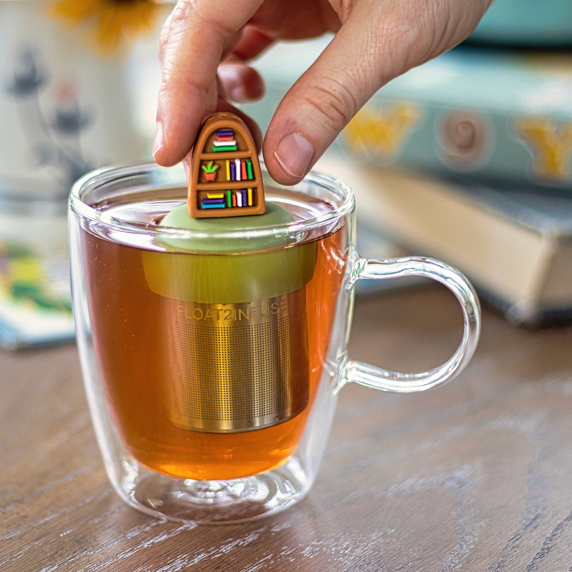 brown bookshelf with books and plants  tea infuser for loose leaf tea  with a fine mesh stainless steel strainer that reads FLOAT2INFUSE on a green base floating in a glass tea mug with books in the background