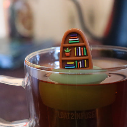 plant bookshelf tea infuser for loose leaf tea with a fine mesh stainless steel tea strainer that reads FLOAT2INFUSE on a green base floating in a glass tea mug with tea kettle in the background