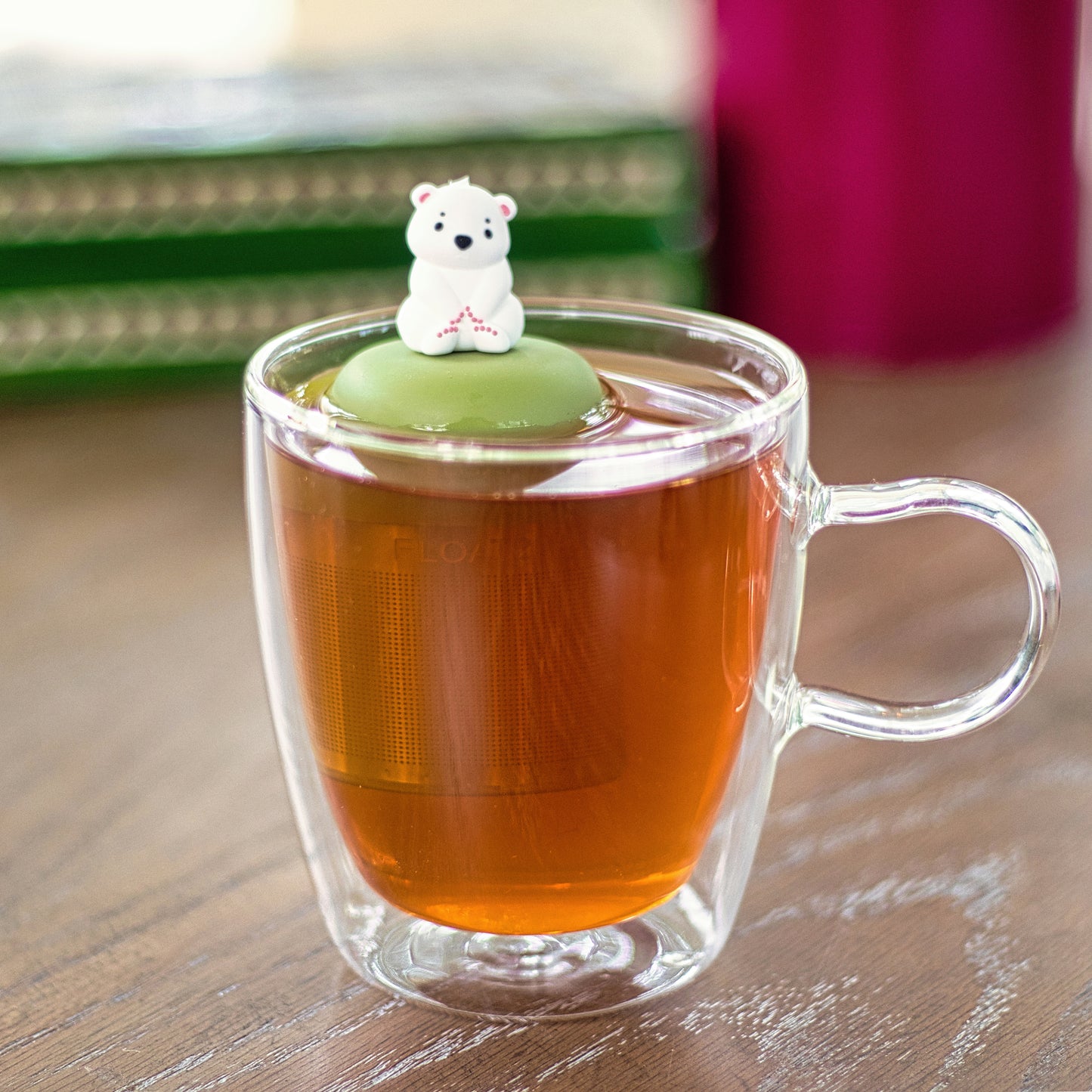 cute white polar bear tea infuser for loose leaf tea with a fine mesh stainless steel tea strainer that reads FLOAT2INFUSE on a green base floating in a glass tea mug 
with books in the background