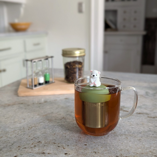 cute white polar bear tea infuser for loose leaf tea with a fine mesh stainless steel tea strainer that reads FLOAT2INFUSE on a green base floating in a glass tea mug 
with loose leaf tea in the background