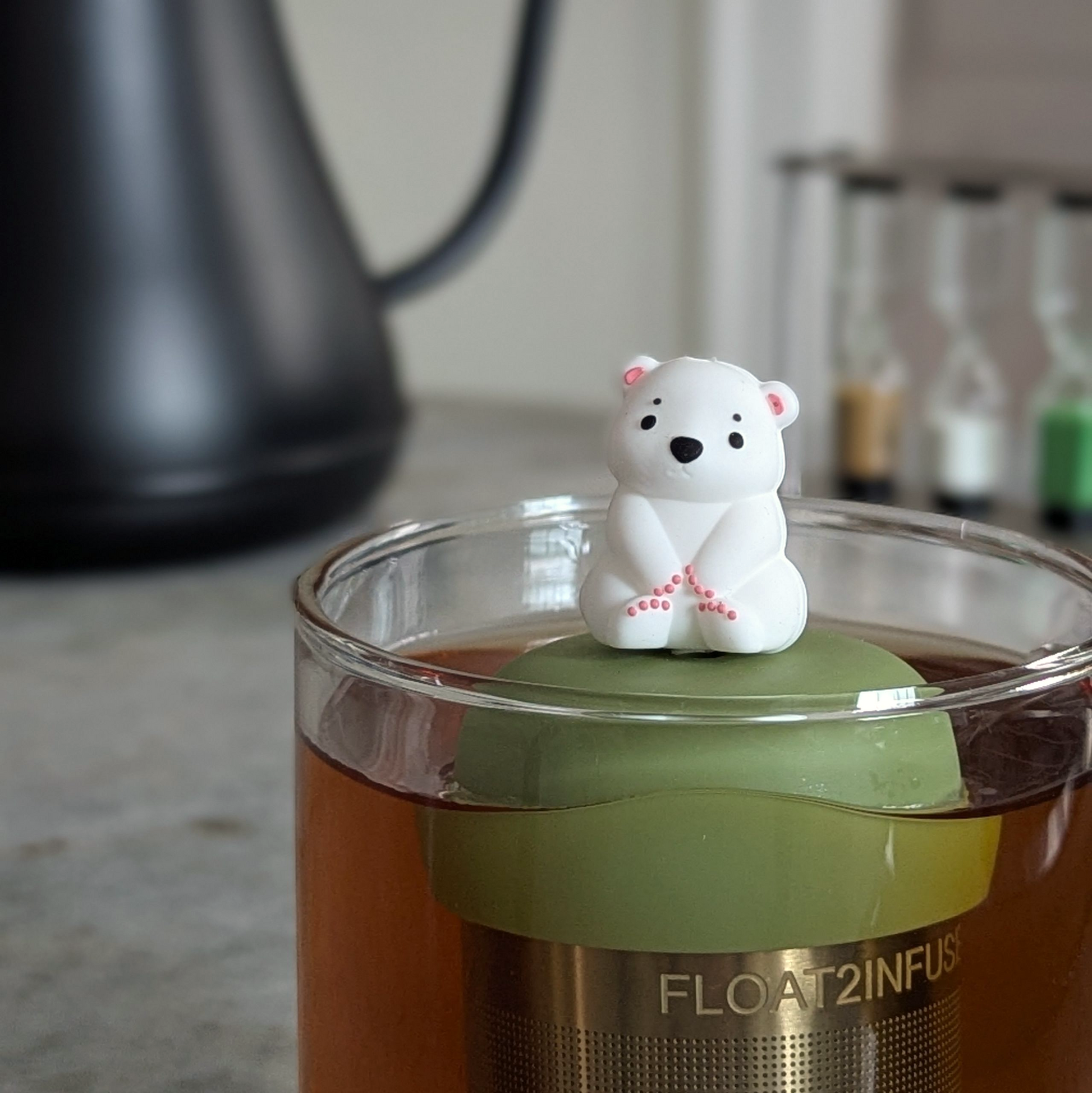 cute white polar bear tea infuser for loose leaf tea with a fine mesh stainless steel tea strainer that reads FLOAT2INFUSE on a green base floating in a glass tea mug 
with a tea kettle in the background