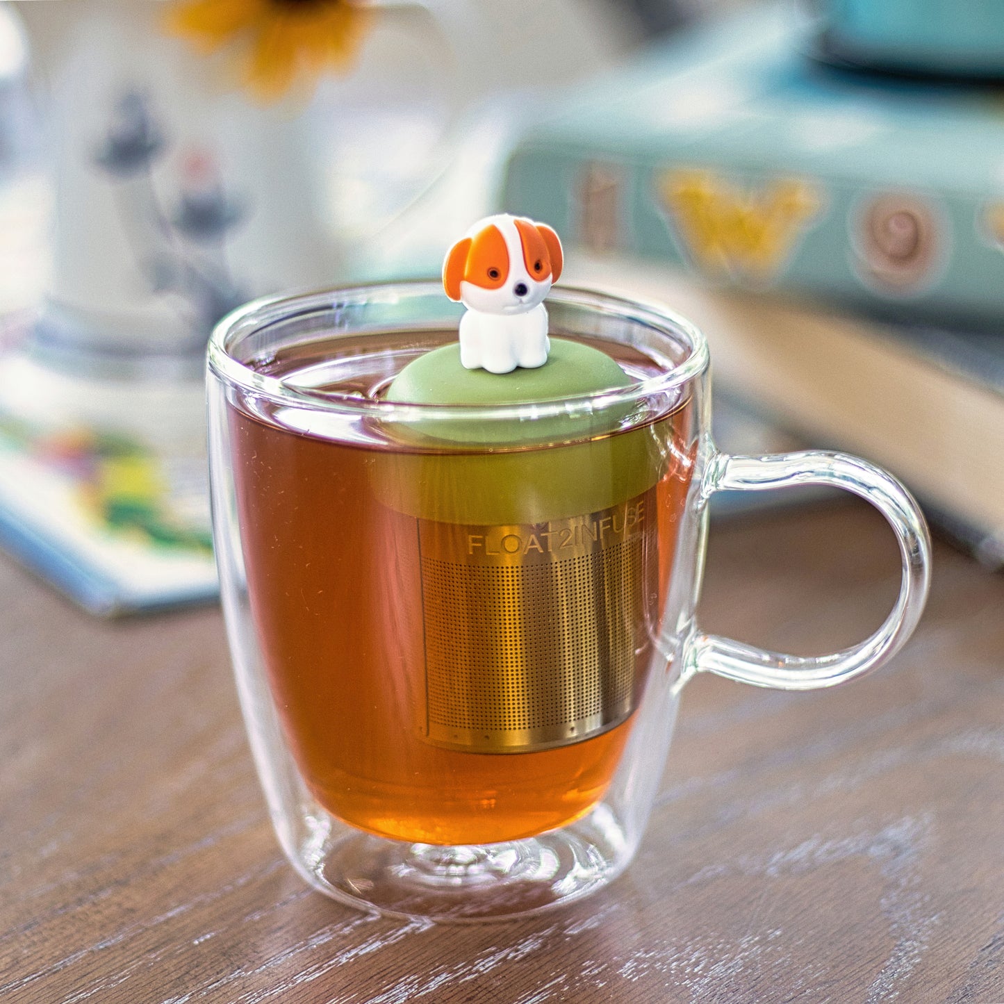 orange and white puppy dog tea infuser for loose leaf tea with a fine mesh stainless steel tea strainer that reads FLOAT2INFUSE on a green base  floating in a glass tea mug with books in the background