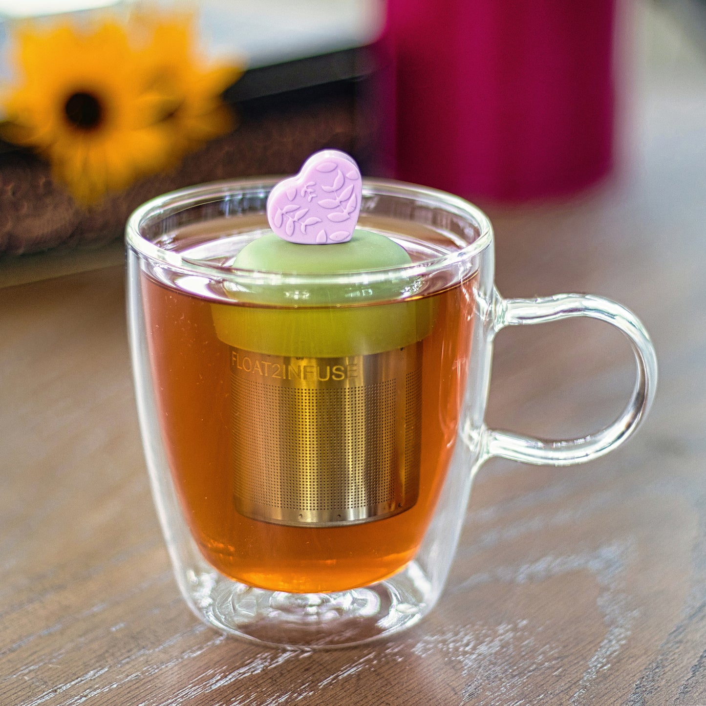 light purple heart with a floral pattern tea infuser for loose leaf tea with a fine mesh stainless steel tea strainer 
that reads FLOAT2INFUSE on a green base  floating in a glass tea mug 
with flowersin the background
