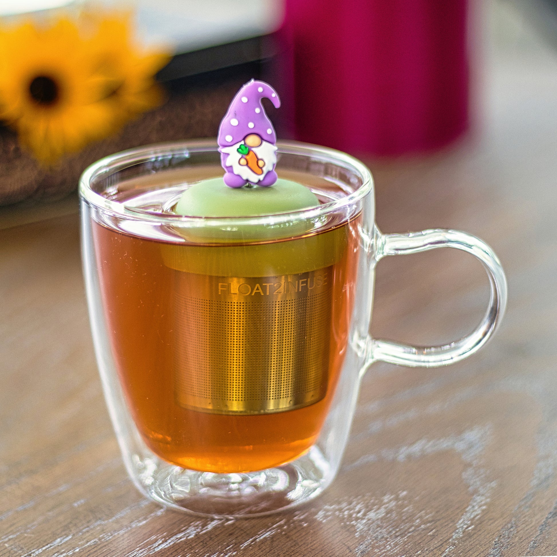 gnome with a long white beard and wearing light purple clothing holding an orange carrot tea infuser for loose leaf tea on a green base with a fine mesh stainless steel strainer that reads FLOAT2INFUSE  floating in a glass tea mug with flowers in the background
