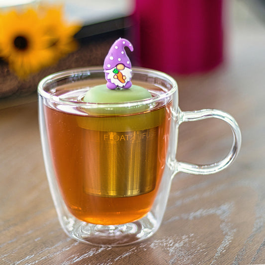 gnome with a long white beard and wearing light purple clothing holding an orange carrot tea infuser for loose leaf tea on a green base with a fine mesh stainless steel strainer that reads FLOAT2INFUSE  floating in a glass tea mug with flowers in the background
