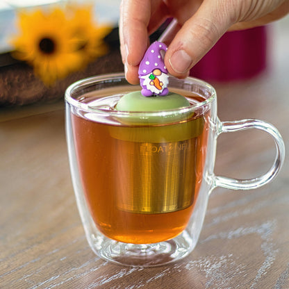 gnome with a long white beard and wearing light purple clothing holding an orange carrot tea infuser for loose leaf tea with a fine mesh stainless steel strainer that reads FLOAT2INFUSE on a green base into a glass tea mug filled with tea with flowers in the background
